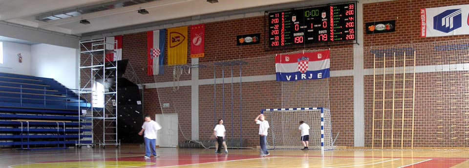 Dvorana s hrvatskim zastavama i natpisom grada, semafor prikazuje vrijeme i rezultat, djeca igraju rukomet